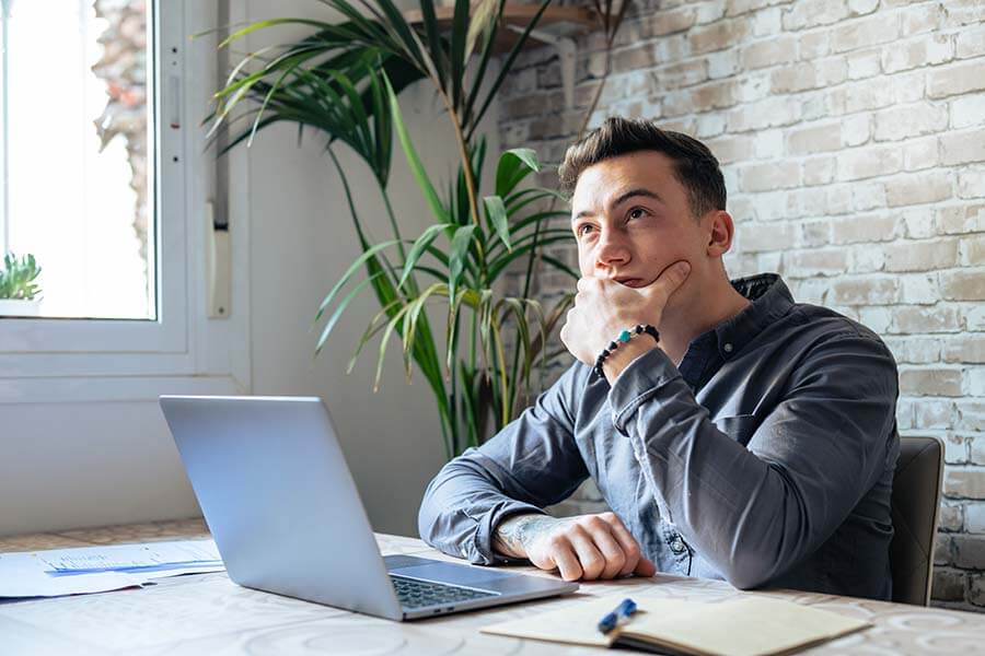 A younger man with his hand on his chin thinks about how to improve his credit score to buy a house.